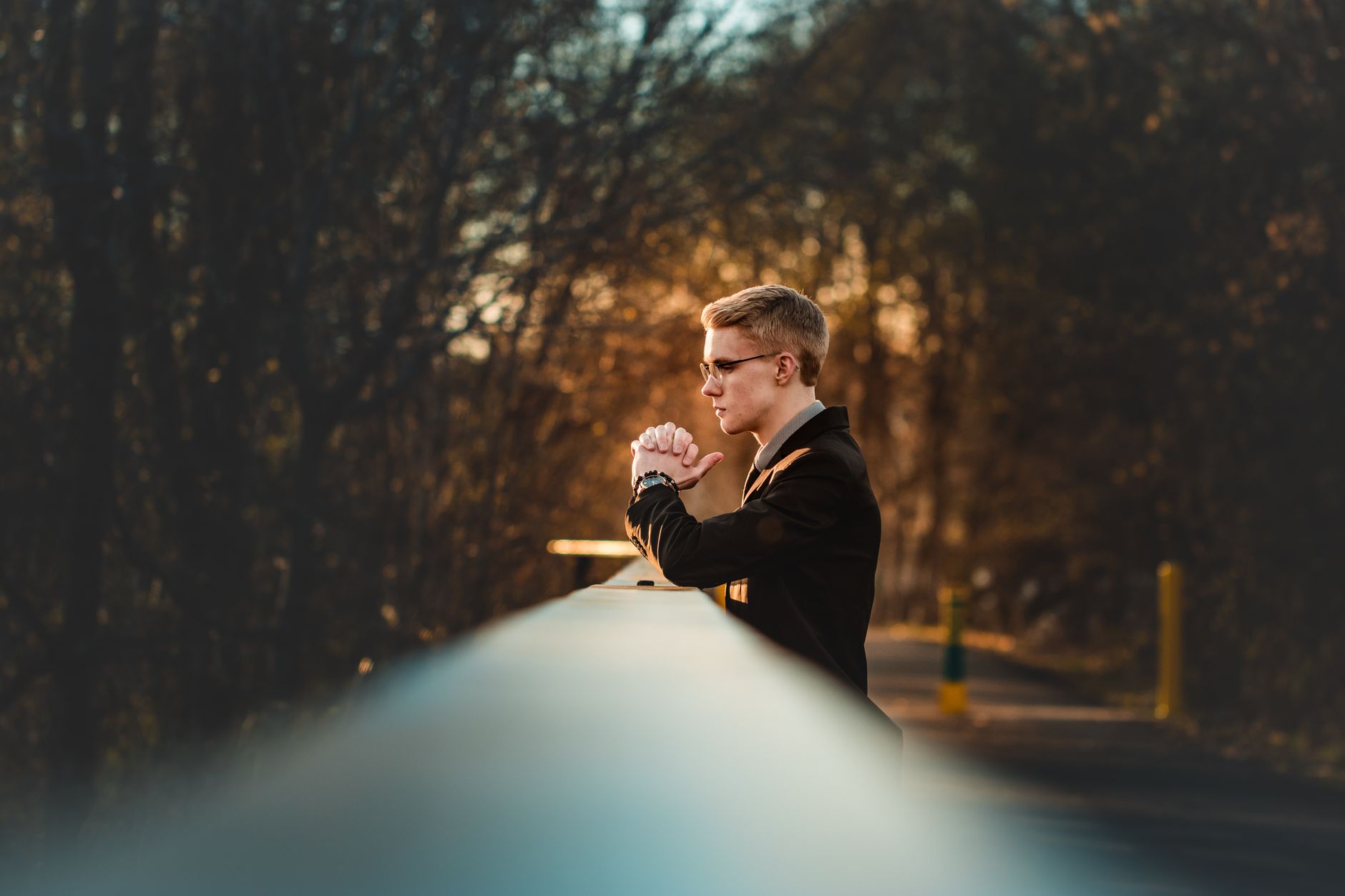 A man in a suit gazes thoughtfully from a bridge during a golden sunset in a park.