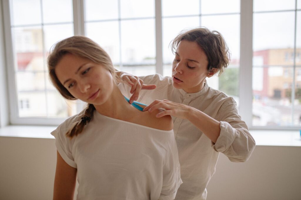 Female therapist applies kinesiology tape to woman's shoulder in healthcare setting.
