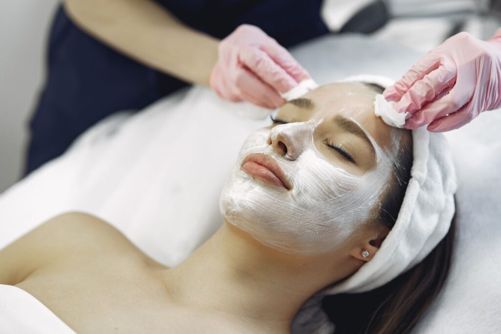 From above anonymous medical specialist in uniform and gloves wiping cream mask off face of relaxed woman during skincare …