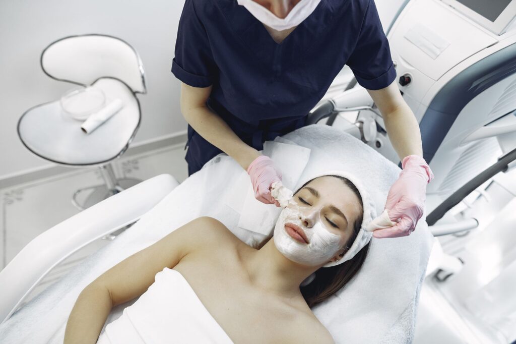 From above of dermatologist in medical coat and latex gloves doing face cleansing and wiping mask from face of female lyin…